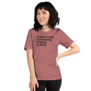 May include: A dusky pink t-shirt featuring the text "Christ-loving, God-fearing, praying mama" in black. The shirt has a crew neck and short sleeves.