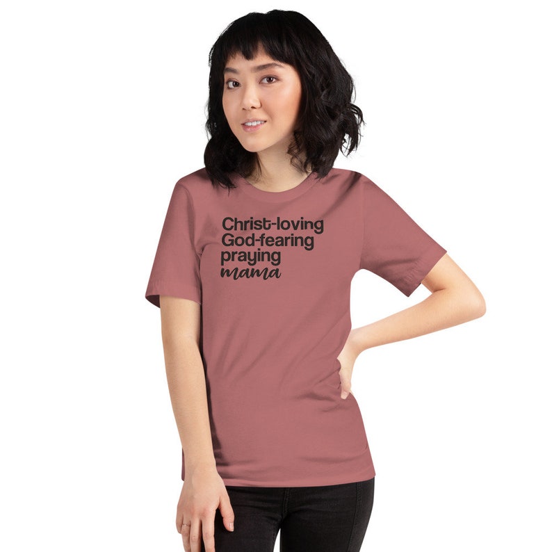 May include: A dusky pink t-shirt featuring the text "Christ-loving, God-fearing, praying mama" in black. The shirt has a crew neck and short sleeves.