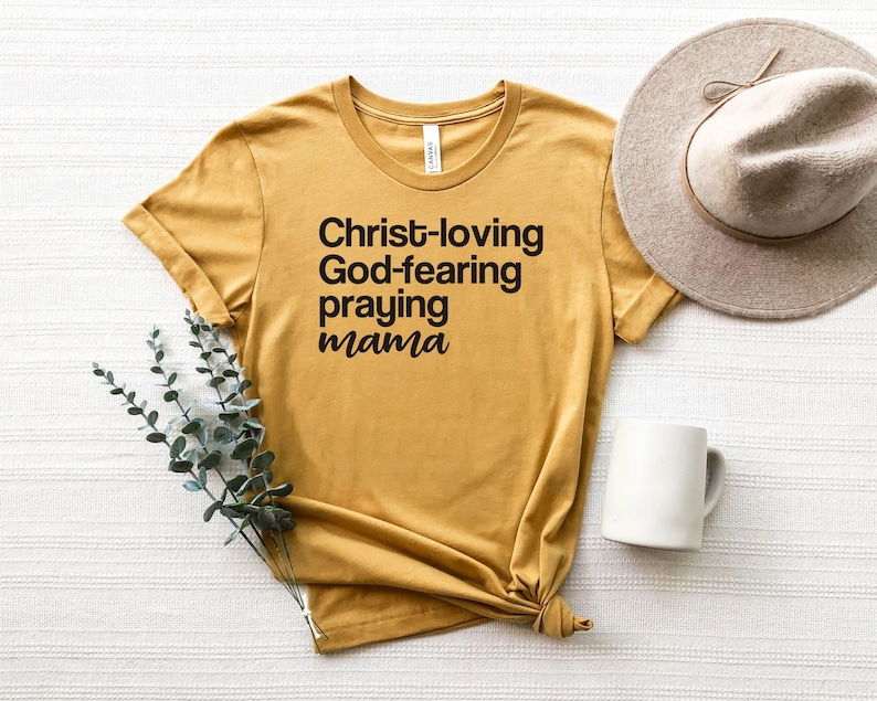 May include: Mustard yellow t-shirt with the words "Christ-loving God-fearing praying mama" in black. A beige hat and white mug are next to the shirt. A sprig of green leaves is placed near the shirt.
