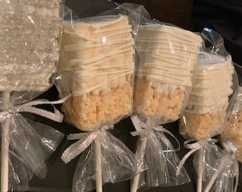 May include: A close-up of six white chocolate rice krispie treats on sticks, individually wrapped in clear plastic bags with silver ribbon bows. The treats are topped with white chocolate and some are sprinkled with white sugar crystals.