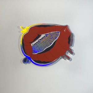 May include: A holographic sticker featuring a stylized mouth with red lips and white teeth. The inside of the mouth has the word "DREAMS" in a holographic design. The sticker has a silver outline and a dripping effect.