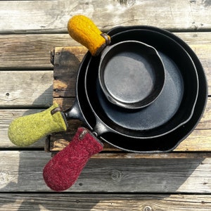 May include: Three cast iron skillets with handles wrapped in colourful wool pot holders. The pot holders are yellow, green and burgundy.