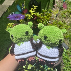 May include: Two small, handmade crochet plush toys featuring round, bright green heads with small ears and black plastic eyes. Each figure wears a white crocheted tunic with a dark brown trim at the hem and dark brown sleeves and feet.