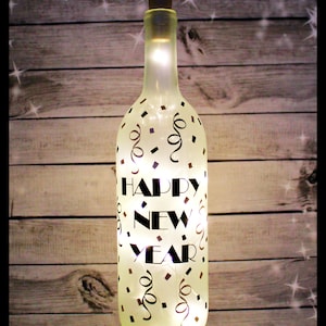 May include: A decorative wine bottle with the words "HAPPY NEW YEAR" illuminated in white. The bottle is decorated with black confetti and swirls. A brown cork sits at the top.