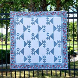 May include: A square quilt with a white background and blue geometric patterns. The quilt features a border with red flowers and blue stripes. The quilt is hanging on a black metal fence.