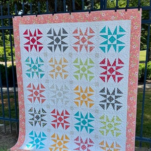 May include: A colorful handmade quilt with a white background and star patterns in various colors, including red, teal, and yellow. The quilt is bordered with a floral pattern and is displayed outdoors.