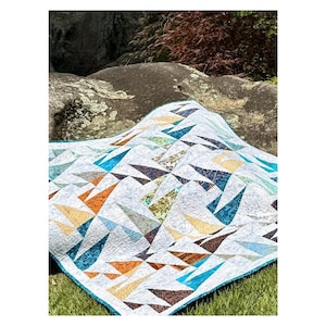May include: A white quilt with a geometric pattern of colorful triangles in shades of blue, orange, brown, and green. The quilt is placed on a rock and grass background, showcasing its design and texture. The quilt has a teal border.
