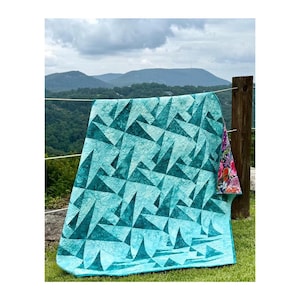 May include: A turquoise quilt with a geometric triangle pattern is draped over a wooden fence. The quilt's reverse side features a floral print. The quilt is displayed outdoors with a scenic mountain backdrop.