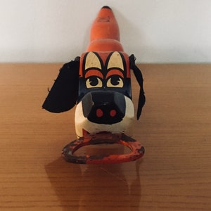 Puede incluir: Un abridor de botellas de juguete de madera vintage en forma de perro con orejas negras, detalles pintados en naranja y negro y una cara blanca con ojos negros y nariz roja. La boca del perro está abierta y tiene un anillo de metal para abrir botellas.