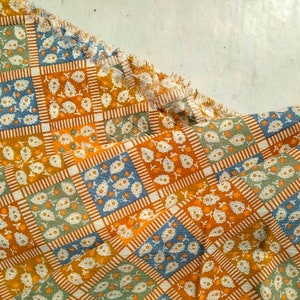 Peut inclure: Gros plan sur un tissu à motifs avec un motif carré répétitif. Chaque carré présente un motif floral blanc sur des fonds bleus, verts et orange. Le tissu a un bord effiloché, suggérant qu'il pourrait s'agir d'un textile pour l'artisanat ou la décoration intérieure.
