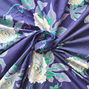 May include: A close-up of a navy blue fabric with a floral pattern. Large white and light blue flowers with yellow centers are scattered across the fabric, along with green and lavender leaves and vines. The fabric is folded, creating shadows and depth.