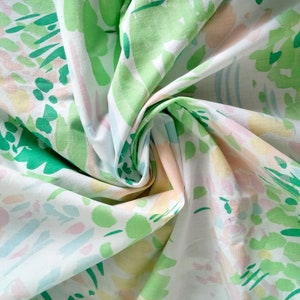 May include: A white fabric featuring an abstract pattern of brushstrokes in shades of light green, dark green, soft pink, and pale yellow. The material is gathered in the center, creating folds and shadows that highlight the printed design.