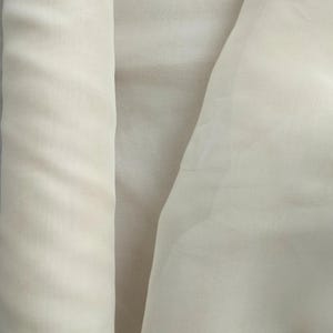 Könnte beinhalten: Nahaufnahme eines cremefarbenen Stoffes, möglicherweise Seide oder Chiffon, mit einer weichen, fließenden Textur. Der Stoff ist gefaltet und drapiert, wodurch Schatten und Lichtreflexe entstehen, die seine zarte Beschaffenheit hervorheben. Die Farbe ist ein blasser, cremiger Farbton.
