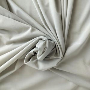 Peut inclure: Un tissu gris clair uni avec une texture lisse et mate. Le matériau est drapé en plis doux et fluides qui créent des ombres et des reflets profonds sur la surface, soulignant la couleur uniforme et le tissage régulier du textile.