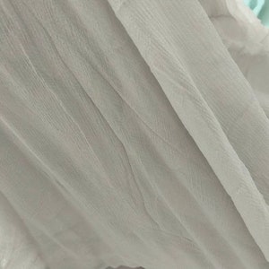 May include: A close-up of a white sheer fabric with a textured surface. The fabric is folded and draped, creating soft folds and wrinkles.