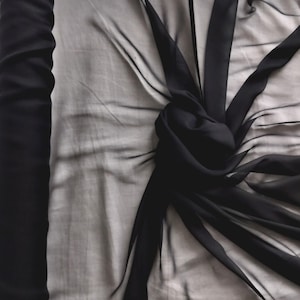 Peut inclure: Tissu noir transparent, peut-être de la mousseline ou de la soie, drapé et noué, créant un effet texturé. Un rouleau du même tissu est visible à gauche. La transparence et la couleur sombre du tissu suggèrent une utilisation pour l'habillement ou la décoration.
