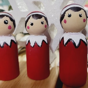 May include: Three wooden elf dolls with painted faces and red and white elf costumes. The dolls are standing on a white surface.