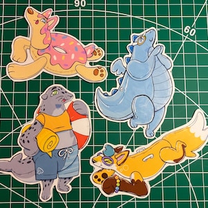 May include: Four cartoon stickers on a green cutting mat. The stickers feature anthropomorphic animals: a turtle with a donut float, a blue dinosaur, a seal with a life preserver, and a fox with sunglasses.