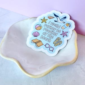 May include: A die-cut sticker with a light blue background featuring the text I WOULD RATHER BE READING AT THE BEACH surrounded by illustrations of a yellow starfish, seashell, beach ball, seagull, bookmark, flip-flops, book, and pink glasses.