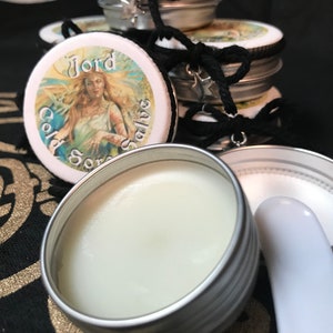 Può includere: Una scatola d'argento con un coperchio bianco che contiene una pomata bianca. Il coperchio ha un'etichetta colorata con l'immagine di una donna e il testo "Jörd Sore Salve".