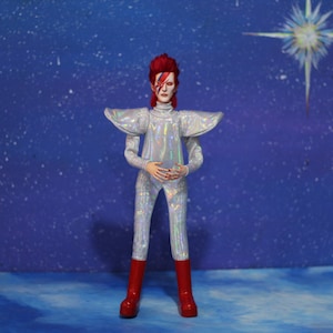 May include: A silver jumpsuit with a red and white lightning bolt design on the face. The jumpsuit has a large collar and is worn by a figure with red hair. The figure is standing in front of a blue background with white stars.