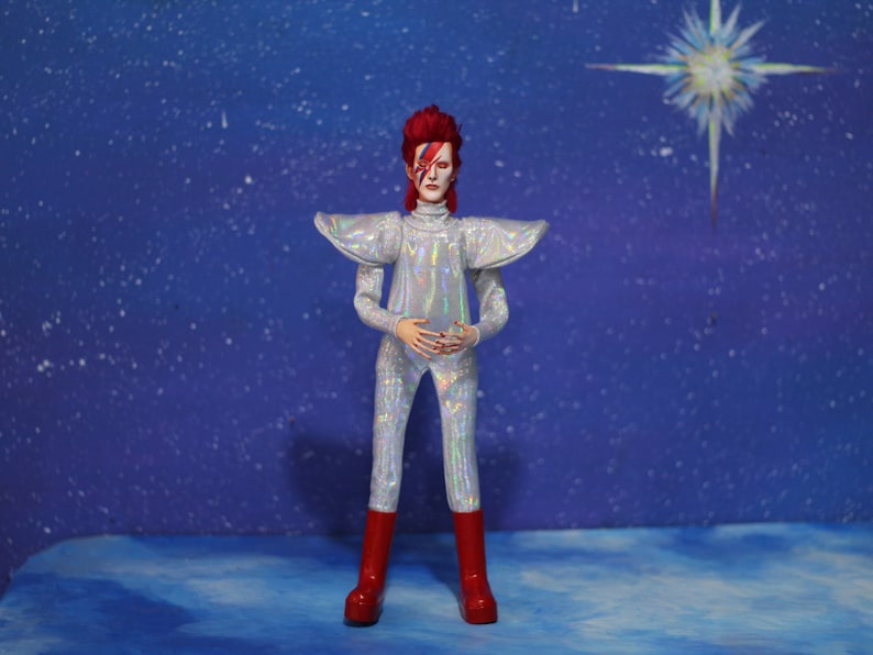 May include: A silver jumpsuit with a red and white lightning bolt design on the face. The jumpsuit has a large collar and is worn by a figure with red hair. The figure is standing in front of a blue background with white stars.