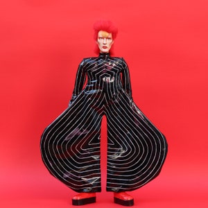 Könnte beinhalten: Puppe in einem schwarz-weiß gestreiften Overall mit ausgestellten Beinen und roten Plateauschuhen. Die Puppe hat rotes Haar und steht vor einem roten Hintergrund.
