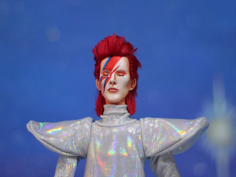 May include: A red-haired doll with a white face and blue and red makeup. The doll is wearing a silver sequined outfit with large, puffed shoulders.