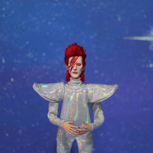 May include: A figure wearing a silver, iridescent jumpsuit with large shoulder pads and a red wig. The figure has blue and red makeup on their face. The background is a blue, sparkly backdrop.