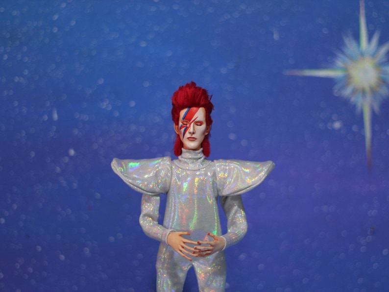 May include: A figure wearing a silver, iridescent jumpsuit with large shoulder pads and a red wig. The figure has blue and red makeup on their face. The background is a blue, sparkly backdrop.