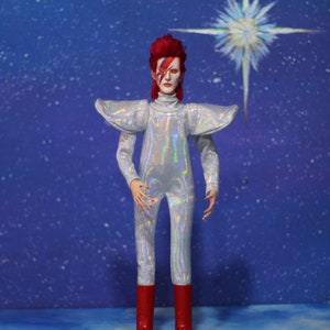 May include: A silver and iridescent jumpsuit with a red and white lightning bolt design on the face. The figure is wearing red boots and has red hair. The background is a blue sky with a white star.
