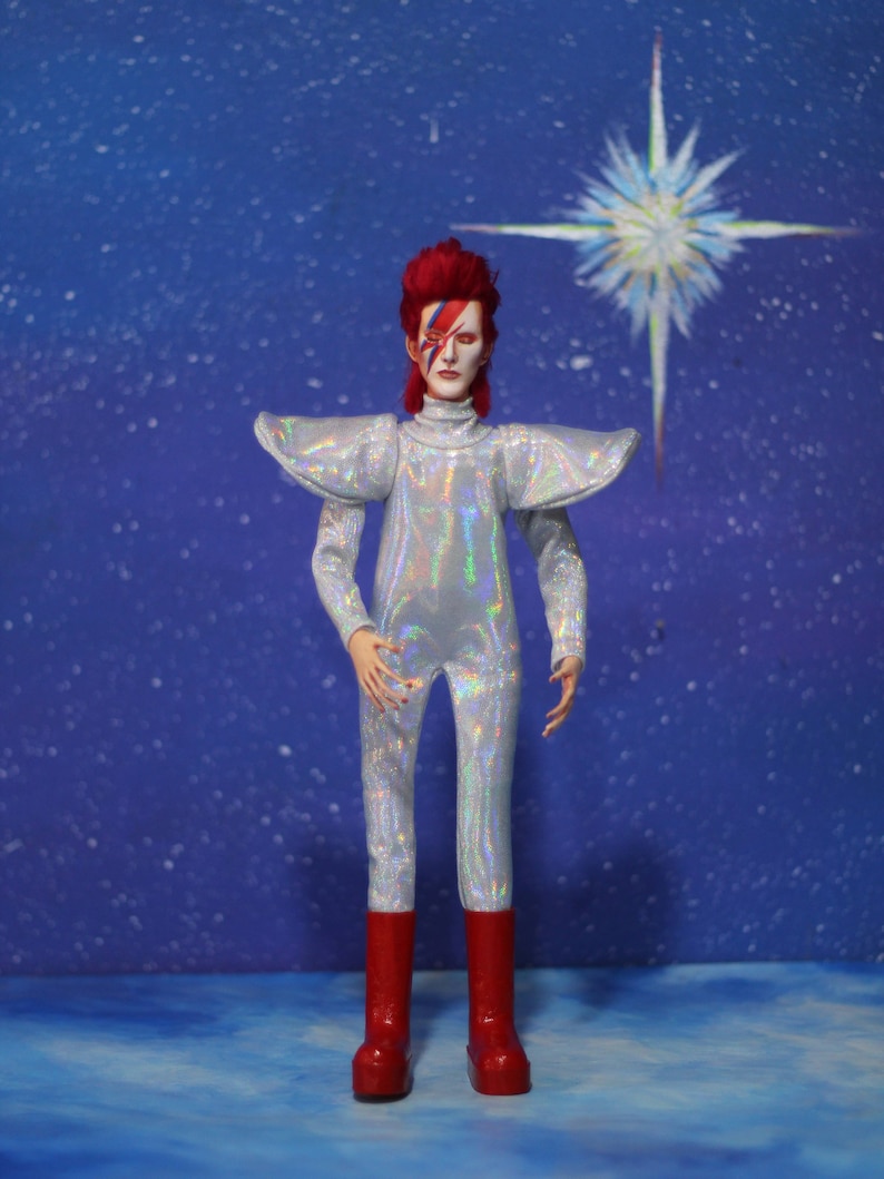 May include: A silver and iridescent jumpsuit with a red and white lightning bolt design on the face. The figure is wearing red boots and has red hair. The background is a blue sky with a white star.