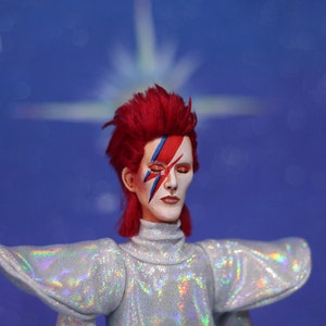 May include: A doll with red hair, a white face and blue and red makeup in the shape of a lightning bolt. The doll is wearing a silver sequined outfit with large, flared sleeves.
