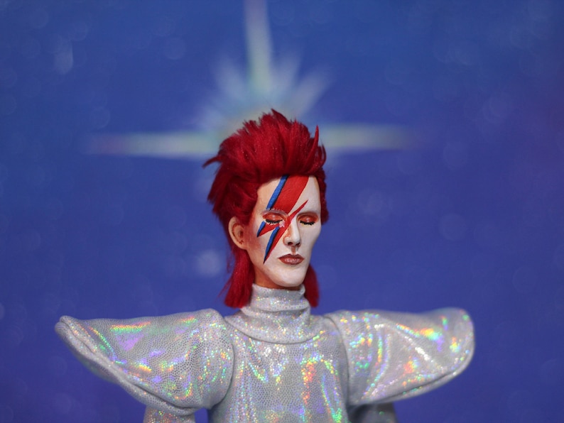 May include: A doll with red hair, a white face and blue and red makeup in the shape of a lightning bolt. The doll is wearing a silver sequined outfit with large, flared sleeves.