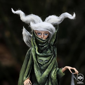 May include: A handcrafted figurine featuring a figure with long, wavy white hair and blue eyes, wearing a draped olive green garment. The fabric includes a painted face detail on the chest area. The figure has visible hands and holds a small gray cloth.
