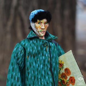 May include: A doll dressed as Vincent van Gogh, wearing a teal coat and a blue hat with black trim. The doll holds a painting of sunflowers. The doll's face is painted with a likeness of the artist.