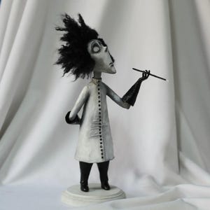 Puede incluir: Una escultura caprichosa en escala de grises de un personaje con cabello negro salvaje, que lleva un abrigo blanco largo con botones negros, guantes y botas negras. La figura sostiene un boquilla y se encuentra sobre una base redonda blanca. La obra de arte tiene un diseño estilizado único.