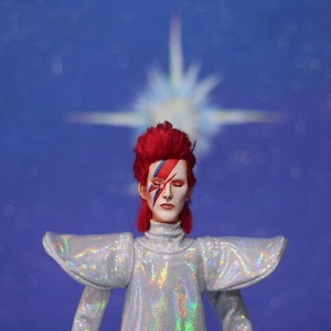 May include: A red-haired doll with a white and blue face paint design, wearing a silver sequined outfit, standing in front of a blue background with a blurred white star.