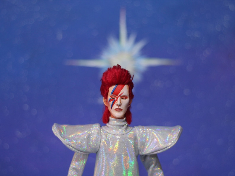 May include: A red-haired doll with a white and blue face paint design, wearing a silver sequined outfit, standing in front of a blue background with a blurred white star.