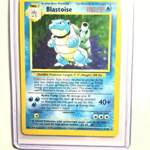 May include: A blue and green Blastoise Pokemon card from the Pokemon Trading Card Game. The card is from the 1999 Wizards set and is number 2/102. The card features Blastoise, a water-type Pokemon, with 100 HP. The card also features the text "Evolves from Wartortle" and "Put Blastoise on the Stage I card".
