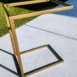 May include: A gold-colored, C-shaped side table with a rectangular top and base, and a vertical support. The table is made of metal and has a modern design. The table is approximately 24 inches tall.