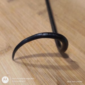 May include: A black metal hook with a curved, tapered end. The object features a smooth, dark finish and a rounded, solid construction that bends into a hook shape, showing a consistent texture throughout the visible portion of the metal piece.