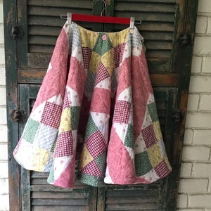 May include: A full-length skirt constructed from quilted fabric featuring a patchwork design. The pattern consists of alternating squares in shades of pink, yellow, green, and white with small floral and checkered prints. It hangs from a red hanger.