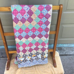 May include: A patchwork quilt featuring a diamond-patterned design composed of numerous small fabric squares in various colors including pink, blue, green, yellow, and black with floral and geometric prints, draped over a wooden rack.