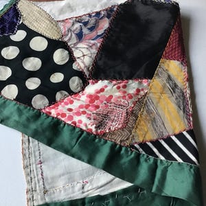 May include: A patchwork textile with a variety of fabrics, including black with white polka dots, floral prints, and striped patterns. The edges are finished with a green satin border. The stitching is visible, adding to the handmade aesthetic.