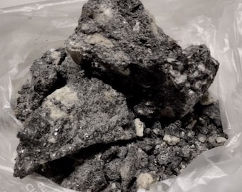 May include: A collection of rough, dark gray rocks with white and silver flecks, presented in a clear plastic bag. The rocks have a coarse, textured appearance, suggesting a natural mineral formation. The overall color palette is muted, with variations in gray and white.