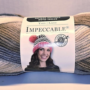 May include: A skein of Impeccable yarn in shades of gray, white, and blue. The label features a woman wearing a pink and black striped hat. The yarn is labeled "Impeccable" and "Yarn | Laine".