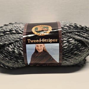 May include: A skein of Lion Brand Yarn, Tweed Stripes, in shades of gray and white. The yarn is wrapped with a label featuring the brand logo and the product name. The label also includes a photo of a woman.