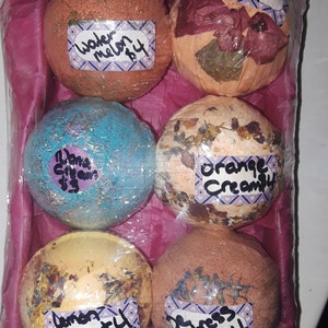 Könnte beinhalten: Sechs Badebomben in einem rosa-weißen Geschenkkorb. Die Badebomben haben verschiedene Farben und Düfte, darunter Wassermelone, Orangencreme und Lavendel-Eukalyptus.