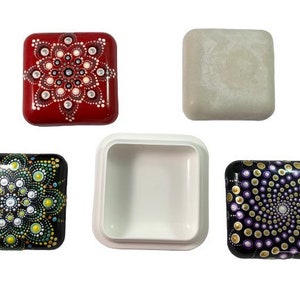 Puede incluir: Cuatro cajas cuadradas con diferentes diseños. Una caja es roja con un diseño de mandala blanco, una es negra con un diseño de mandala verde y blanco, una es morada con un diseño de mandala blanco y dorado, y una es blanca lisa.